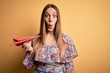 © Krakenimages.com - Young blonde woman holding paper plane over yellow isolated background scared in shock with a surprise face, afraid and excited with fear expression