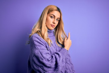 Wall Mural - Young beautiful blonde woman wearing casual turtleneck sweater over purple background Pointing with hand finger to the side showing advertisement, serious and calm face