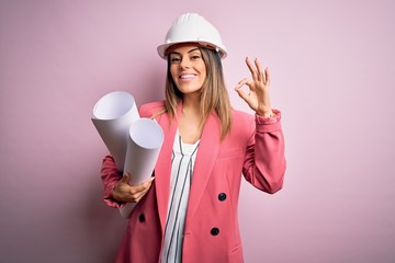 Wall Mural - Young beautiful brunette architect woman wearing safety helmet holding blueprints doing ok sign with fingers, excellent symbol