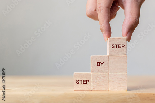 Fotomural Hand arranging wood block stacking as step stair on top with word Step By Step