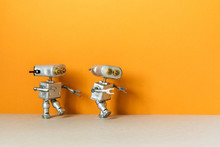 Two Metal Silver Robots Are Walking. Simplified Symbolic Toy Robotic Characters On An Orange Background. Empty Space For Text.