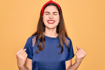 Poster - Young beautiful brunette woman wearing red lips over yellow background very happy and excited doing winner gesture with arms raised, smiling and screaming for success. Celebration concept.