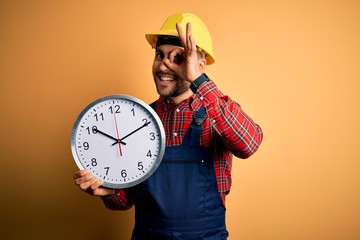 Canvas Print - Young builder man wearing safety helmet holding big clock over yellow background with happy face smiling doing ok sign with hand on eye looking through fingers