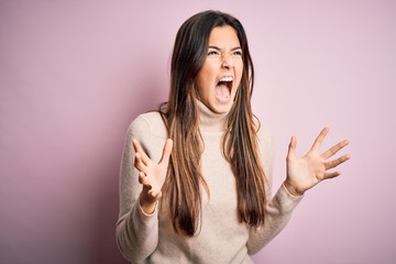 Poster - Young beautiful girl wearing casual turtleneck sweater standing over isolated pink background crazy and mad shouting and yelling with aggressive expression and arms raised. Frustration concept.