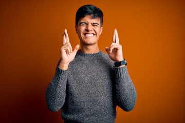 Poster - Young handsome man wearing casual sweater standing over isolated orange background gesturing finger crossed smiling with hope and eyes closed. Luck and superstitious concept.