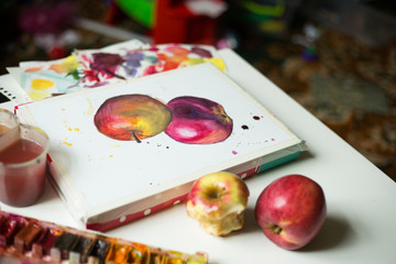 On a white table is a drawing, two red apples, watercolor paint, water in a glass