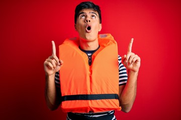 Sticker - Young handsome man wearing orange safety life jacket over isolated red background amazed and surprised looking up and pointing with fingers and raised arms.