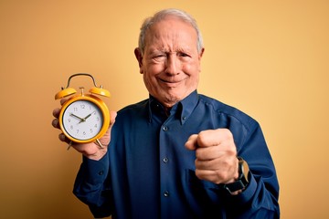 Senior grey haired man holding vintage alarm clock over yellow background annoyed and frustrated shouting with anger, crazy and yelling with raised hand, anger concept