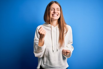 Young beautiful redhead sporty woman wearing sweatshirt over isolated blue background very happy and excited doing winner gesture with arms raised, smiling and screaming for success. Celebration