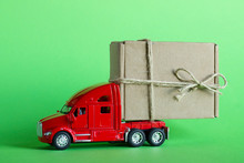 Red Toy Car Truck Delivers A Parcel In A Craft Cardboard Box On A Pastel Green Background. Delivery Of Goods, Gifts Or Donation Concept.