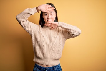 Young beautiful asian woman wearing casual sweater over yellow isolated background Smiling cheerful playing peek a boo with hands showing face. Surprised and exited