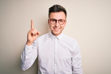 Sticker - Young handsome business mas wearing glasses and elegant shirt over isolated background showing and pointing up with finger number one while smiling confident and happy.