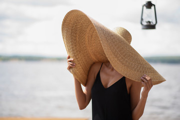 A young woman on a beach wearing a giant straw hat and a lighting is close to her 