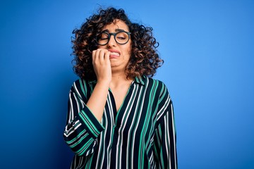 Sticker - Young beautiful curly arab woman wearing striped shirt and glasses over blue background looking stressed and nervous with hands on mouth biting nails. Anxiety problem.