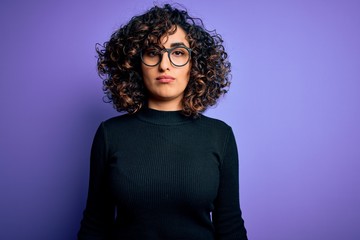 Poster - Young beautiful curly arab woman wearing casual sweater and glasses over purple background with serious expression on face. Simple and natural looking at the camera.