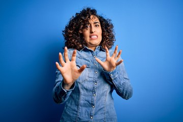 Sticker - Young beautiful curly arab woman wearing casual denim shirt standing over blue background disgusted expression, displeased and fearful doing disgust face because aversion reaction. With hands raised