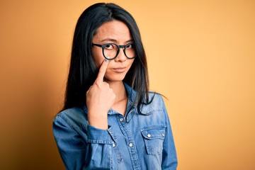 Wall Mural - Young beautiful chinese woman wearing casual denim shirt over isolated yellow background Pointing to the eye watching you gesture, suspicious expression