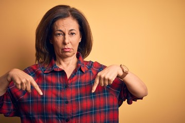 Poster - Middle age beautiful woman wearing casual shirt standing over isolated yellow background Pointing down looking sad and upset, indicating direction with fingers, unhappy and depressed.