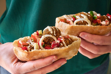 Woman hands hold a two half of pita bread sandwich with fresh authentic falafels balls inside and chopped salad and drizzle of tahini sauce on top, close-up of chickpea falafel in gluten-free pita