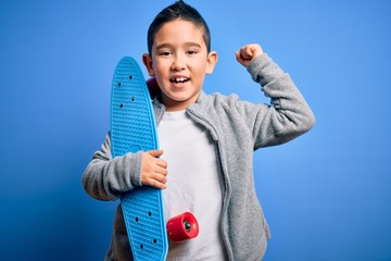 Poster - Young little boy kid skateboarder holding modern skateboard over blue isolated background screaming proud and celebrating victory and success very excited, cheering emotion