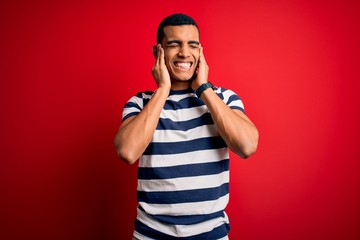 Handsome african american man wearing casual striped t-shirt standing over red background covering ears with fingers with annoyed expression for the noise of loud music. Deaf concept.