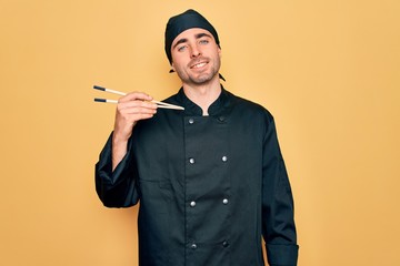 Wall Mural - Young handsome cooker man with blue eyes wearing uniform and hat using chopsticks with a happy face standing and smiling with a confident smile showing teeth