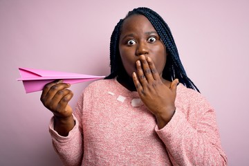 Sticker - Young african american plus size woman with braids holding pink paper airplane cover mouth with hand shocked with shame for mistake, expression of fear, scared in silence, secret concept