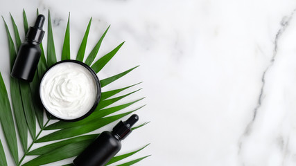 Flat lay jar of organic cream, palm leaf and dropper bottles on marble background. Top view SPA natural cosmetics, skincare concept