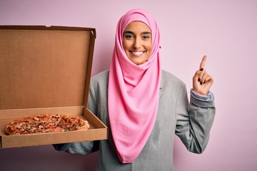 Young beautiful arab woman wearing pink muslim hijab holding delivery box with Italian pizza surprised with an idea or question pointing finger with happy face, number one