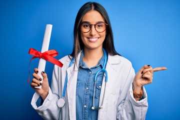 Wall Mural - Young beautiful brunette doctor woman wearing glasses and coat holding diploma degree very happy pointing with hand and finger to the side