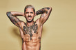 © Cien X Cien Studio/ADDICTIVE STOCK - Handsome sexy attractive muscular male with various tattoos on naked torso and arms looking at camera making faces with mouth while standing against beige background