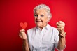 Senior beautiful woman holding paper heart standing over isolated red background screaming proud and celebrating victory and success very excited, cheering emotion