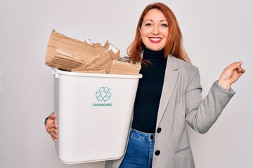 Poster - Young beautiful redhead woman recycling holding trash can with cardboard to recycle very happy pointing with hand and finger to the side