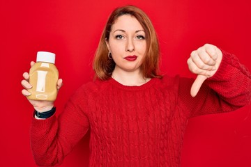 Sticker - Beautiful redhead woman holding bottle of mustard sauce condiment over red background with angry face, negative sign showing dislike with thumbs down, rejection concept