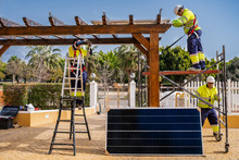 Group Of Workers In Uniform And Helmets Installing Photovoltaic Panels On Roof Of Wooden Construction Near House