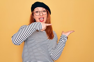 Wall Mural - Beautiful redhead woman wearing striped t-shirt and french beret over yellow background amazed and smiling to the camera while presenting with hand and pointing with finger.