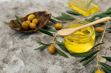 From Above Of Glass Jar With Organic Olive Oil With Wooden Spoon Placed On Shabby Gray Table Near Fresh Green Olives And Tree Branches