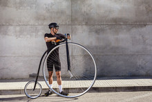 Calm Determined Man In Black Activewear And Helmet Looking Away While Standing On Asphalt Road And Leaning On High Wheel Bicycle Against Concrete Wall In Sunny Day