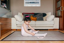 Side View Of Cute Girl In Leotard And Tights Sitting On Floor Near Sofa And Putting On Dance Shoes Before Ballet Rehearsal At Home