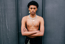 Alluring Young Black Teenage Man With Naked Torso And Neck Chains Holding Flower In Mouth And Looking At Camera While Standing Against Gray Wall