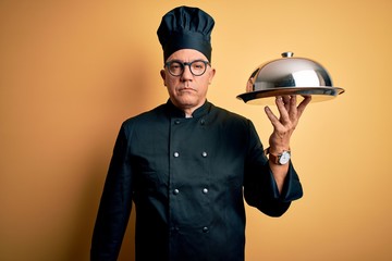 Poster - Middle age handsome grey-haired waiter man wearing cooker uniform and hat holding tray Relaxed with serious expression on face. Simple and natural looking at the camera.