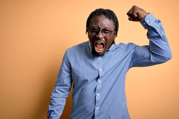 Canvas Print - Young handsome african american man wearing shirt and glasses over yellow background angry and mad raising fist frustrated and furious while shouting with anger. Rage and aggressive concept.