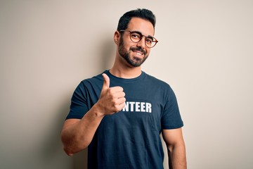 Handsome man with beard wearing t-shirt with volunteer message over white background doing happy thumbs up gesture with hand. Approving expression looking at the camera showing success.