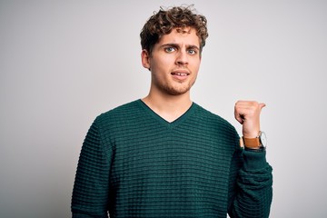 Young blond handsome man with curly hair wearing green sweater over white background smiling with happy face looking and pointing to the side with thumb up.