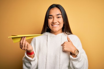 Canvas Print - Young beautiful asian woman holding paper airplane standing over isolated yellow background with surprise face pointing finger to himself