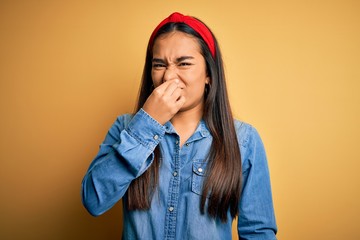 Poster - Young beautiful asian woman wearing casual denim shirt and diadem over yellow background smelling something stinky and disgusting, intolerable smell, holding breath with fingers on nose. Bad smell