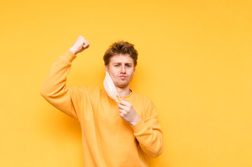 A funny young man takes off his meditation mask and looks at the camera with a skeptical face and a raised hand out of joy, isolated on a yellow background. Coronavirus pandemic. Quarantine. covid-19.