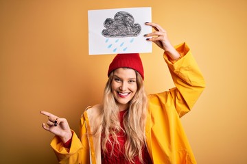 Sticker - Young beautiful blonde woman wearing raincoat and wool cap holding banner with cloud image very happy pointing with hand and finger to the side