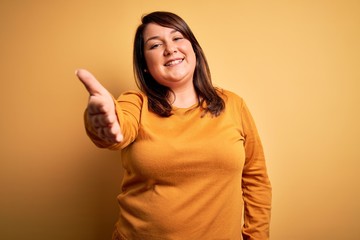 Wall Mural - Beautiful brunette plus size woman wearing casual sweater over isolated yellow background smiling friendly offering handshake as greeting and welcoming. Successful business.