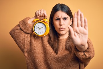 Poster - Young beautiful woman holding alarm clock standing over isolated orange background with open hand doing stop sign with serious and confident expression, defense gesture